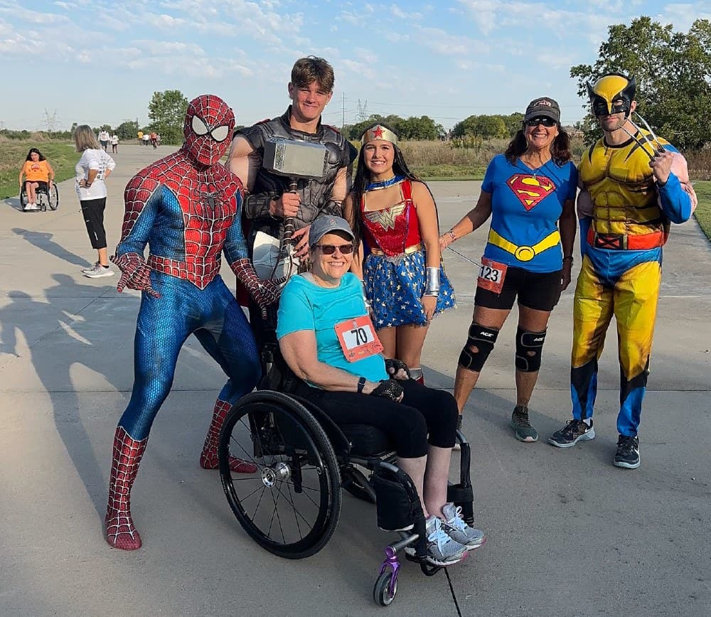 A participant in a wheelchair poses with costumed superheroes and runners at a previous Powered to Move Superhero 5K. Photo courtesy of Powered to Move